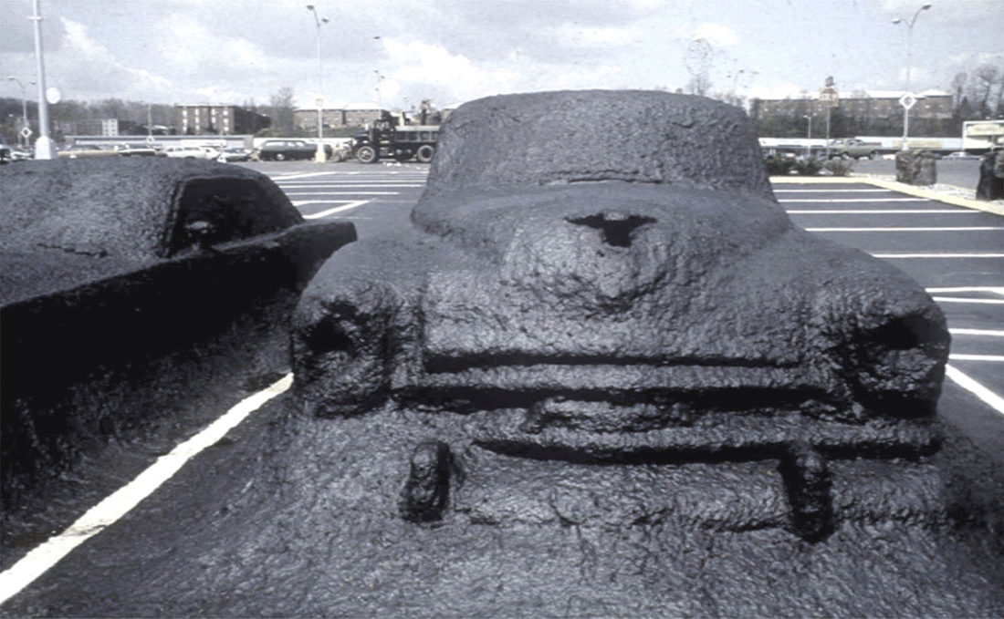 Opera d'arte di James Wines: in un grande parcheggio un'automobile è completamente coperta di asfalto, fusa con la pavimentazione e immobilizzata.