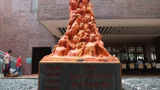The photo shows Jens Galschiøt's sculpture "The Pillar of shame" in front of a brick building. On the basement there are two red writing "The old cannot kill the young" and "THE TIANANMEN MASSACRE". Over the basement the lower part of the sculpture is visbile, with the orange sculpted half-busts of the martyrs