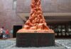 La foto mostra la scultura "The Pillar of Shame" di Jens Galschiøt davanti ad un edificio in mattoni. Della scultura sono mostrati il piedistallo, su cui sono incise le scritte in rosso "The old cannot kill the young" e "THE TIANANMEN MASSACRE" e la parte inferiore in cui sono ben visibili i mezzi busti scolpiti dei martiri dipinti di arancione