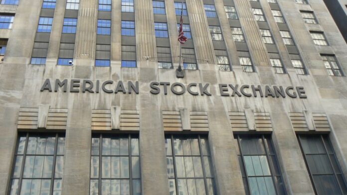 La foto mostra la sede dell'American Stock Exchangge building, un edificio grigio in stile Art decò, pieno di finestree con una bandiera statunitense appessa ad un pinnacolo al centro