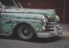 La foto mostra la parte anteriore di un'auto verde anni '50. la vernice è in gran parte sbiadita e la vettura si presenta in generale in condizioni pessime. Sullo sfondo è visibile un garage dal portone arruginito