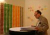 Man with glasses browsing a book. In front of him a drawing hangs on the wall with red, green, orange and brown strips