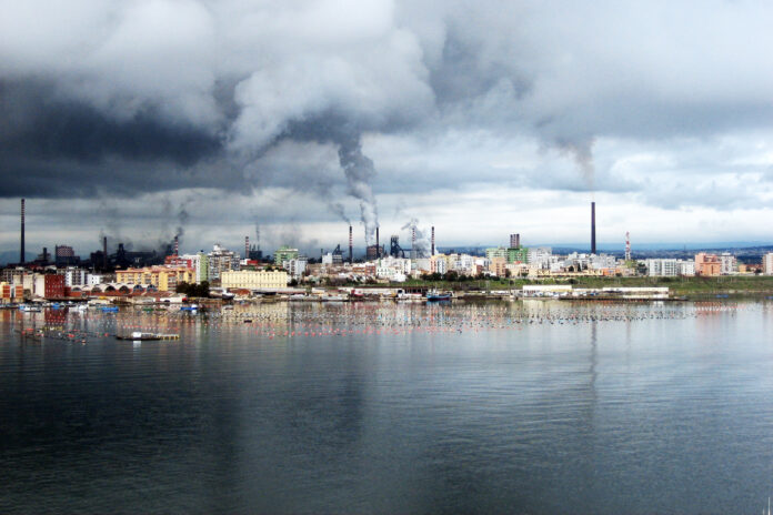 scenario urbano con palazzi e ciminiere di fabbriche affacciato sul mare. Dalle ciminiere fuoriescono fumi che rendono grigio il cielo
