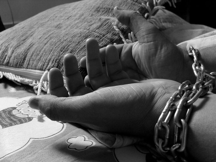 The picture is a black and white photography showing a pair of chained hands near a pillow on a bed cover with infantile decorations