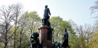 un monumento che raffigura tre personaggi con in basso l'artista Christian Etongo vestito di bianco con una sciarpa rossa