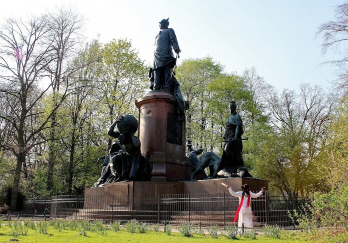 un monumento che raffigura tre personaggi con in basso l'artista Christian Etongo vestito di bianco con una sciarpa rossa
