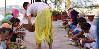 The photo represents an Indian man dressed in white and yellow providing food to a couple of parallel lines of people. The meal consist of a dish with some rice and a iron glass of water