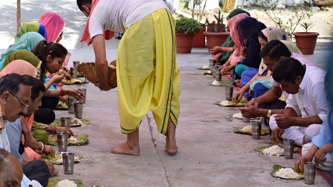 The photo represents an Indian man dressed in white and yellow providing food to a couple of parallel lines of people. The meal consist of a dish with some rice and a iron glass of water