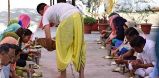 La foto mostra un uomo in abiti indiani gialli e bianchi distribuire cibo a due file di persone disposte parallelamente. il rancio dei presenti è costituito da un piatto di riso ed un bicchiere d'acqua