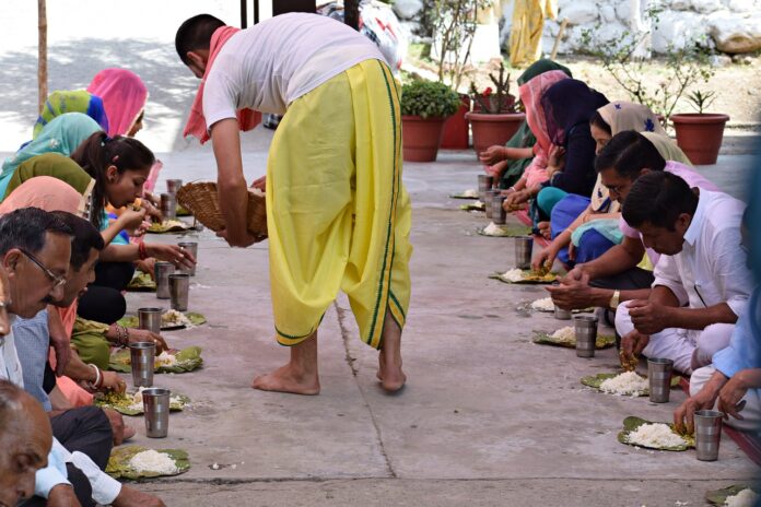 La foto mostra un uomo in abiti indiani gialli e bianchi distribuire cibo a due file di persone disposte parallelamente. il rancio dei presenti è costituito da un piatto di riso ed un bicchiere d'acqua