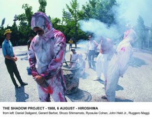 man with a white jumpsuit spotted with red. He keeps head down and is surrounded by men wearing the same jumpsuit, surrounded in turn by smoke. Other people look at the scene