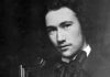 L'immagine è una foto in bianco e nero che mostra André Gide in gioventù- Il giovane, posizionato nella parte destra della fotografia ha capelli di media lunghezza e un accenno di barba. veste una giacca con un fiocchetto e siede su una bella sedia su sfondo scuro
