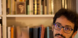 nella foto, sulla destra, si vede il volto di un giovane scrittore con la barba, i baffi e gli occhiali neri; ha gli occhi neri. Sullo sfondo una libreria bianca.