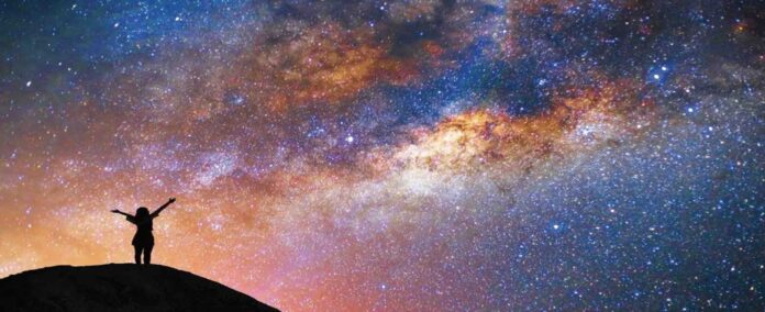 Il principio antropologico di John Barrow cielo stellato invaso in buona parte da una luce gialla. In controluce sagoma di una persona in piedi con le braccia spalancate verso il cielo