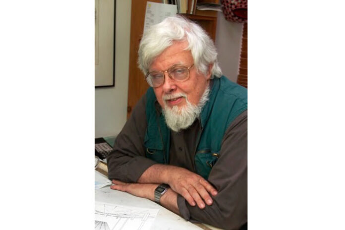 L'immagine mostra James Wines, seduto ad un tavolo con le braccia conserte mentre guarda vrso l'osservatore. Ha capelli, barba e baffi bianchi, porta occhiali e veste una camicia grigia con un gilet verde scuro