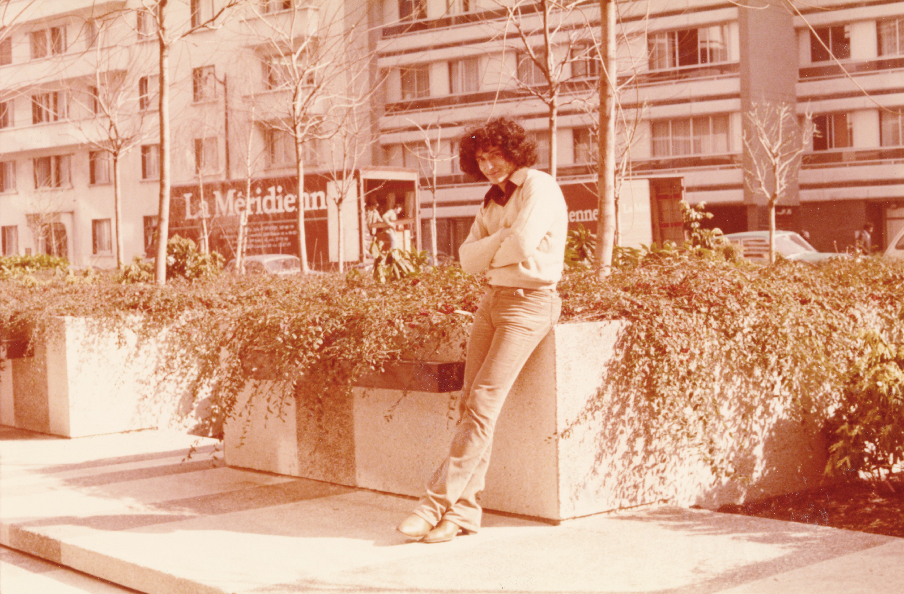 The picture is a sepia photograph showing young Bedri Baykam during his journey to France in 1978. The artist has black curly hair and he's leaning on a flowerbed with some houses in the background