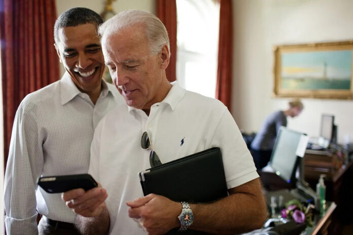 in the colored photo you can see two men, Barack Obama and Joe Biden. Biden wears glasses on the shirt and puts in his hands a tablet and a tape recorder.