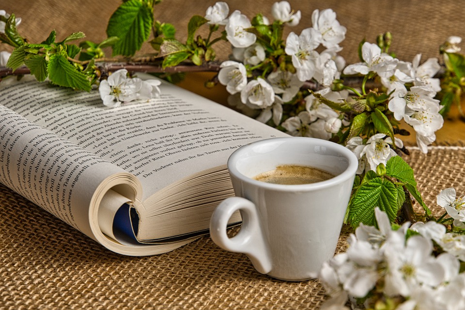 nella foto a colori si vede in primo piano una tazzina con caffè, un libro aperto e un ramo di fiori bianchi su un tavolo
