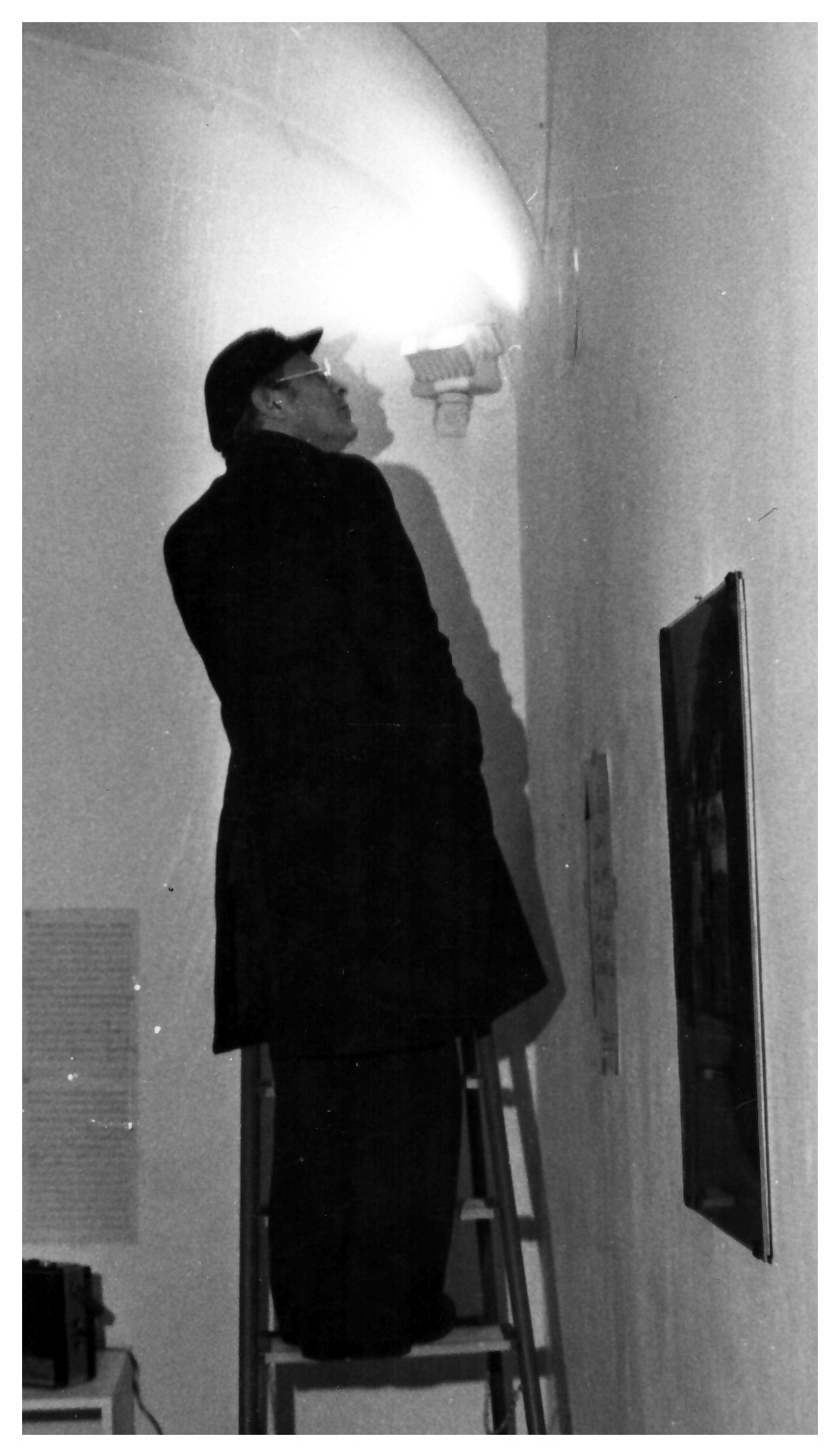 L'immagine mostra Filiberto Menna intento a completare la preparazione della sua mostra alla Galleria Lastaria di Roma. L'uomo, vestito in giacca e pantaloni neri e in piedi su una scala a pioli, intento ad osservare da vicino la luce di un faretto