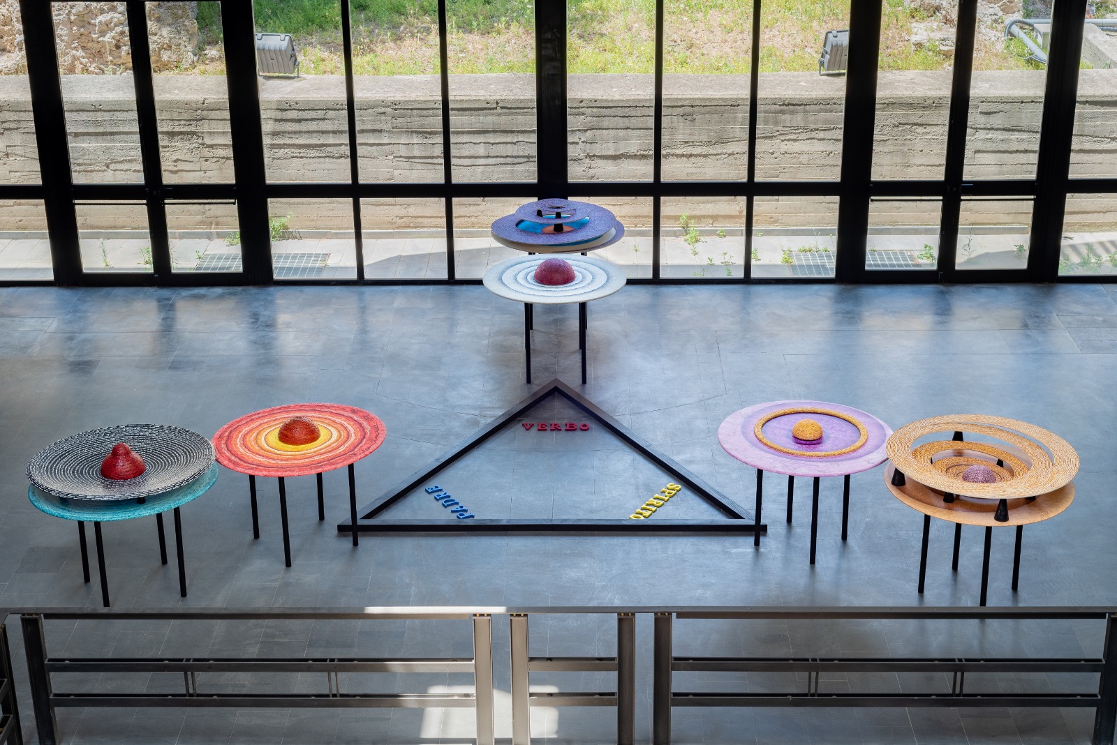 Unpublished visual of the artist Rosario Genovese composed of six tables with different colors and materials placed at the top of a triangle, on the ground, with a black perimeter, inside whose vertices there are the words verb, mother, spirit.