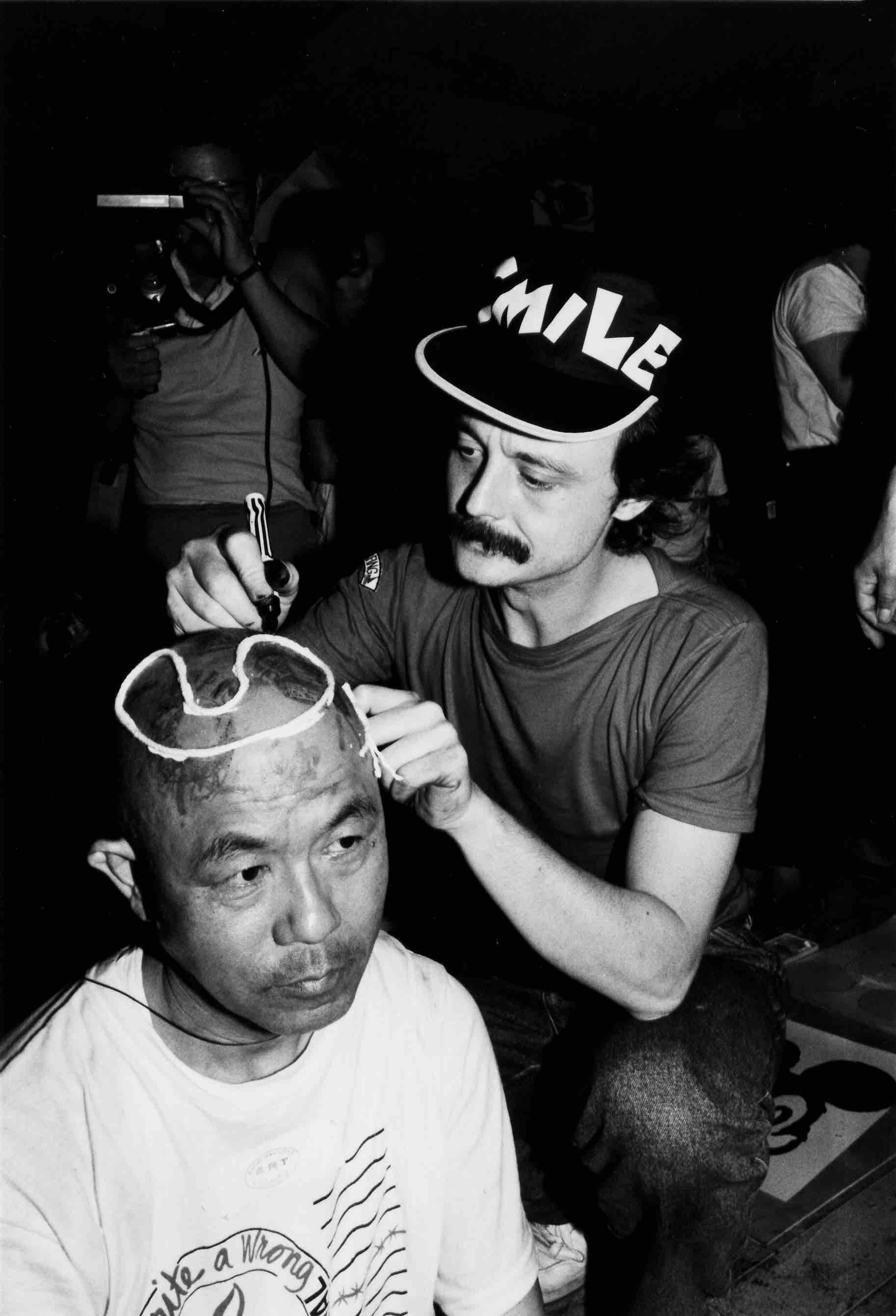 Nella foto: due uomini, uno europeo con un cappello con la scritta smile e uno giapponese che indossa una maglietta con la scritta mail art, sono seduti. Il primo scrive con un pennarello sulla testa del secondo.