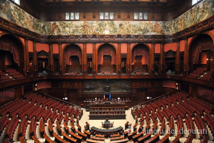 nella foto a colori si vede l'emiciclo della Camera dei Deputati, con, al centro, la postazione in cui siede il presidente della Camera.
