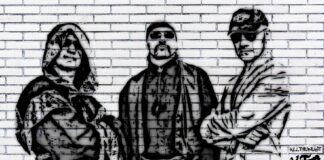 nella foto in bianco e nero si vedono tre uomini dipinti su un muro; indossano occhiali da sole e quello a sinistra un mantello