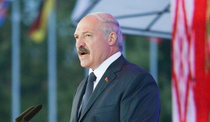 nella foto a colori si vede il dittatore bielorusso Alexander Lukashenko che tiene un discorso di fronte ad una pedana e un microfono; indossa completo scuro e cravatta scura, o blu o nero.