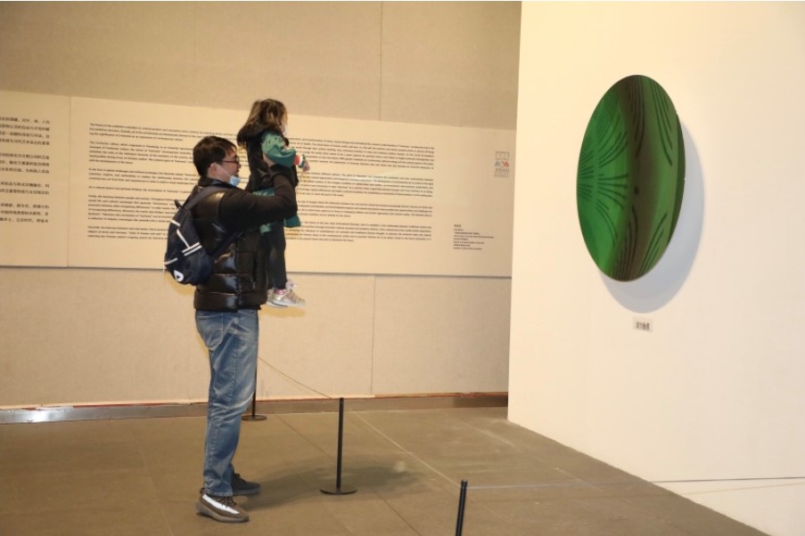 nella foto a colori si vede un'opera d'arte su una parete rotonda e di colore verde, mentre un signore con una bambina in braccio la sta ammirando.
