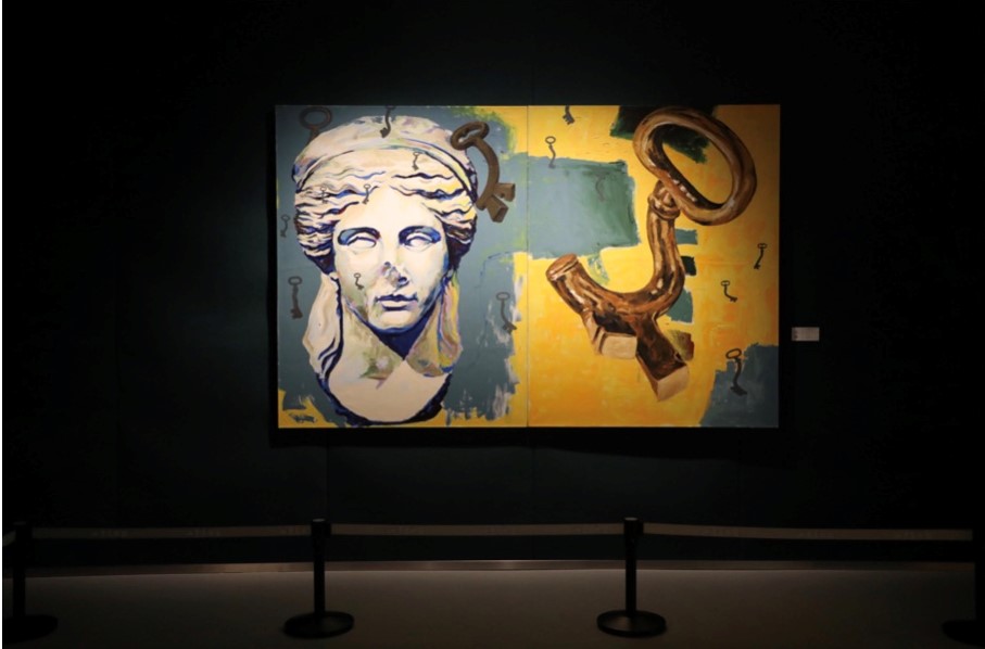 nella foto a colori si vede un'opera d'arte su una parete, composta, a destra, dalla testa di una statua antica e, a sinistra, da una specie di chiave piegata e marrone su sfondo giallo.