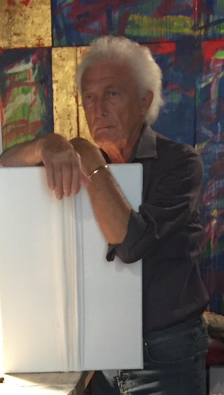 La foto mostra l'artista Giampiero Podestà nel suo studio. Un uomo anziano dai capelli bianchi in camicia nera appoggiato su una tavola di legno bianco. Lo sfondo è occupato da una serie di tele, tendenti principalmente al blu, a parte due completamente dorate