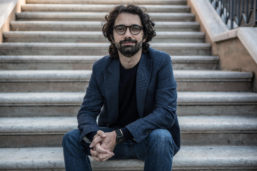 L'immagine è una foto di Andrea de Nicola, uno degli autori del libro Cosa Loro, Cosa Nostra. L'uomo ha capelli neri e lunghi, barba e occhiali. Indossa una giacca di tessuto blu, una maglietta nera e jean. L'uomo è seduto alla base di una scalinata