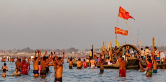 La foto mostra una folla di fedeli Indù immersi nelle acque del Gange. La maggior parte di essi, sono in acqua a torso nudo, mentre sullo sfondo, una barca stracolma di persone e con due bandiere rosse icon alcune scritte in sanscrito attraversa il fiume