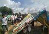 nella foto a colori si vede un campo con tende colorate, forse un campo profughi, con alcune persone di colore in piedi sulla sinistra. A destra, in primo piano, una tenda.