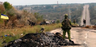 L'immagine mostra una strada nella regione ucraina del Donbass pattugliata da soldati dell'esercito nazionale di Kiev. Un soldato in primo piano è in piedi accanto ad un mucchio di pietre a bordo strada, mentre poco oltre altri soldati occupano l'area attorno ad una macchina ferma. Una bandiera ucraina è appesa ad alcuni arbusti nella parte sinistra dell'immagine