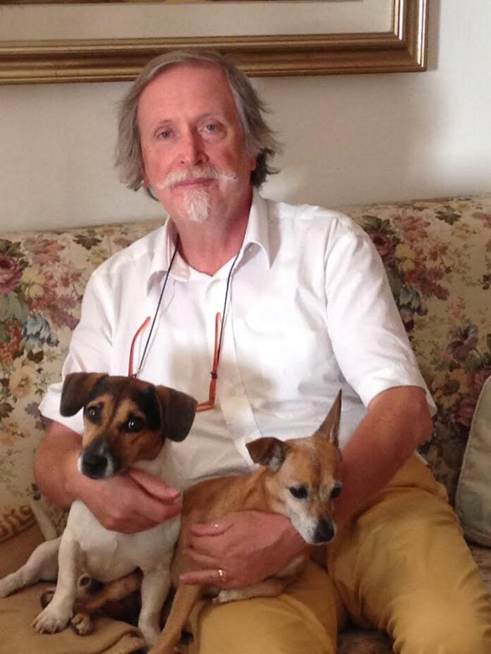 la foto a colori mostra lo scrittore Roberto Barbolini che tiene in braccio due cani; indossa una camicia bianca a maniche corte, occhiali con la catenella e pantaloni beige
