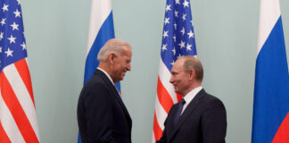 la foto a colori mostra in primo piano il Vicepresidente Usa Joe Biden che stringe la mano al Primo Ministro russo Vladimir Putin; sullo sfondo le bandiere dei due Paesi