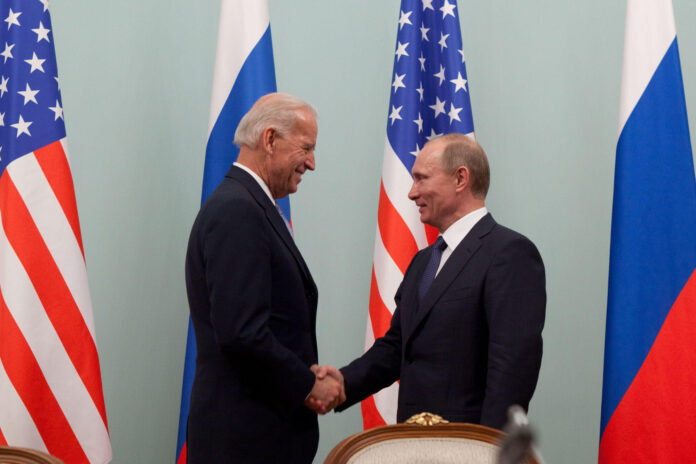 la foto a colori mostra in primo piano il Vicepresidente Usa Joe Biden che stringe la mano al Primo Ministro russo Vladimir Putin; sullo sfondo le bandiere dei due Paesi