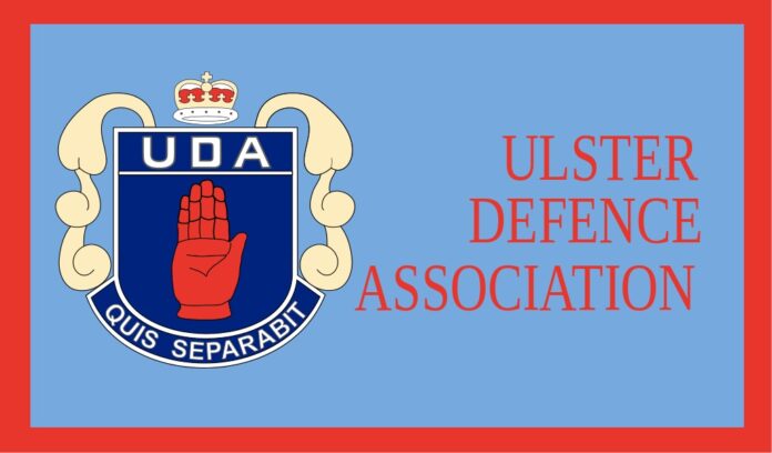 L'immagine mostra il logo della Ulster Defence Association, una mano rossa alzata su uno scudo blu contornato di bianco con una corona e la sigla UDA sopra ed il motto 