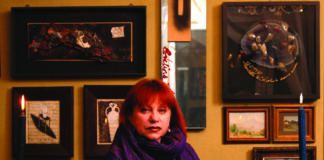 Fotografia che mostra Schatz, artista la cui patria è la Russia, seduta di fronte a un tavolo su cui vi sono due candelabri con candele accese, una mano tesa in avanti, sullo sfondo una parete con opere d'arte appese