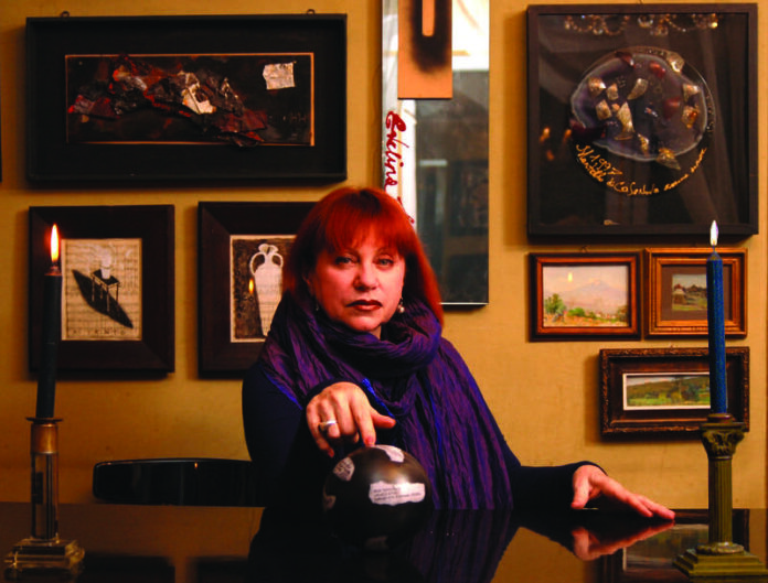 Fotografia che mostra Schatz, artista la cui patria è la Russia, seduta di fronte a un tavolo su cui vi sono due candelabri con candele accese, una mano tesa in avanti, sullo sfondo una parete con opere d'arte appese