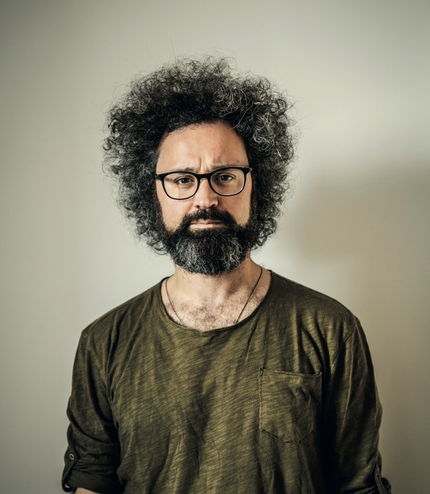 la foto a colori mostra il cantautore Simone Cristicchi che indossa occhiali da vista con la montatura nera, ha i capelli ricci neri leggermente un po' bianchi, porta la barba e i baffi. Infine, indossa un maglione color giallo spento e nero.