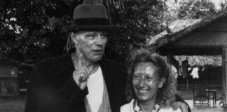 Foto in bianco e nero, un uomo con un cappello sul capo e un cappotto abbraccia amichevolmente una donna sorridente che indossa abiti chiari estivi, in un luogo esterno con molti alberi