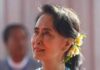 la foto a colori mostra l'attivista birmana Aung San Suu Kyi che porta i capelli castani raccolti in una coda di cavallo fermata da fiori gialli; indossa una collana di perline turchesi e un abito bianco con una balza dello stesso colore della collana