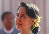 la foto a colori mostra l'attivista birmana Aung San Suu Kyi che porta i capelli castani raccolti in una coda di cavallo fermata da fiori gialli; indossa una collana di perline turchesi e un abito bianco con una balza dello stesso colore della collana