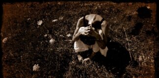 L'immagine è una foto a color seppia che mostra una donna seduta su un prato con qualche fiore qua e la. La donna è inquadrata dall'alto e tiene la testa tra le gambe e le mani sul capo, probabilmente triste o disperata