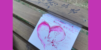 l'immagine mostra un foglio di carta piegato infilato tra le assi di legno di una panchina. Sul foglio è disegnato un cuore con acquarello rosa ed alcune scritte