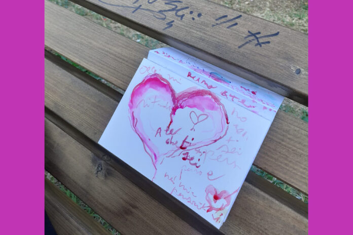 l'immagine mostra un foglio di carta piegato infilato tra le assi di legno di una panchina. Sul foglio è disegnato un cuore con acquarello rosa ed alcune scritte