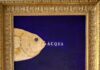 L'immagine mostra una delle opere di Sabatino della serie Pescibarocchi. Un piccolo quadro blu con un pesce giallo emergente dalla parte sinistra dell'immagine con al centro la scritta ACQUA illuminata. L'opera è contornata da una classica cornice in legno