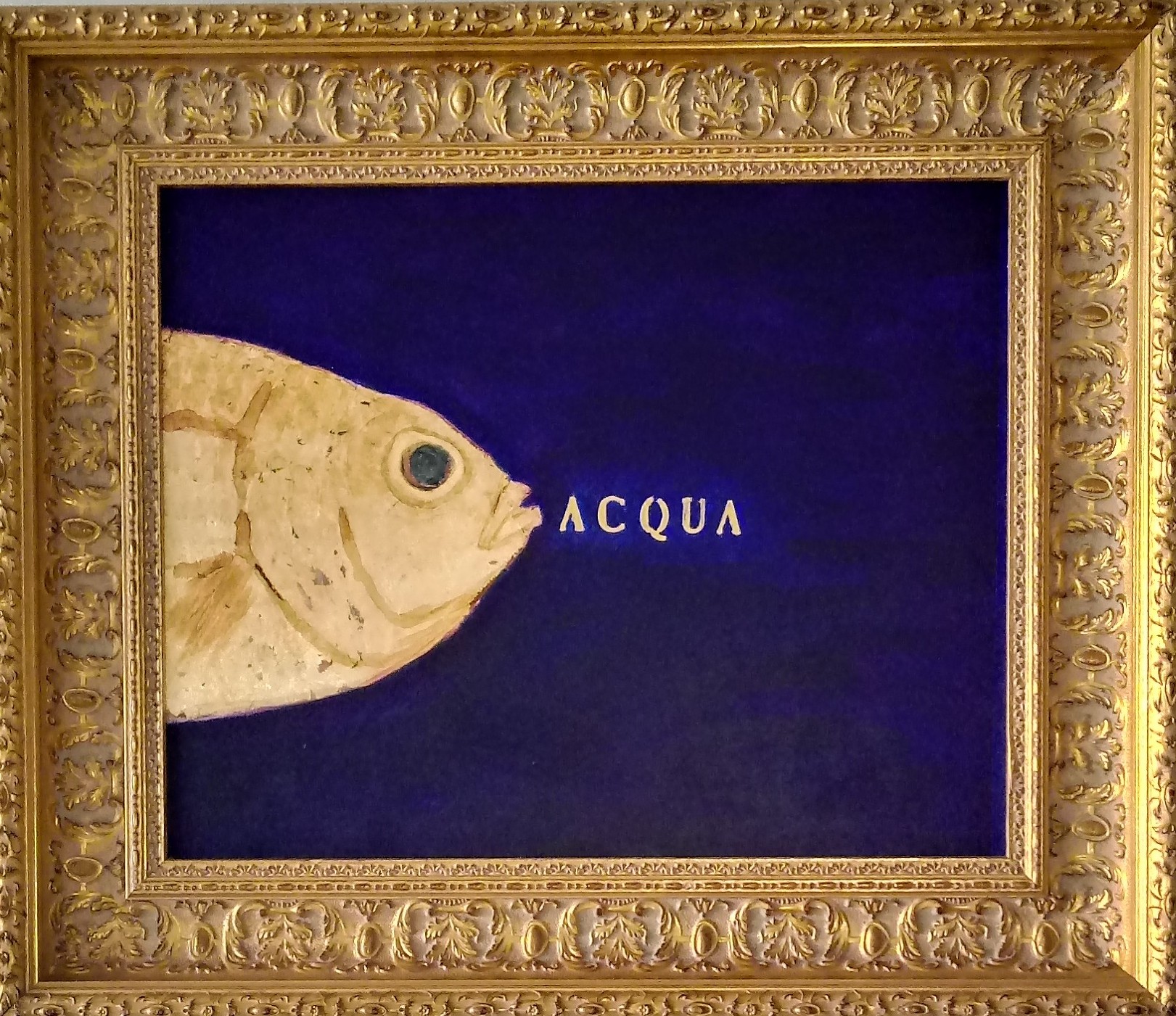 L'immagine mostra una delle opere di Sabatino della serie Pescibarocchi. Un piccolo quadro blu con un pesce giallo emergente dalla parte sinistra dell'immagine con al centro la scritta ACQUA illuminata. L'opera è contornata da una classica cornice in legno